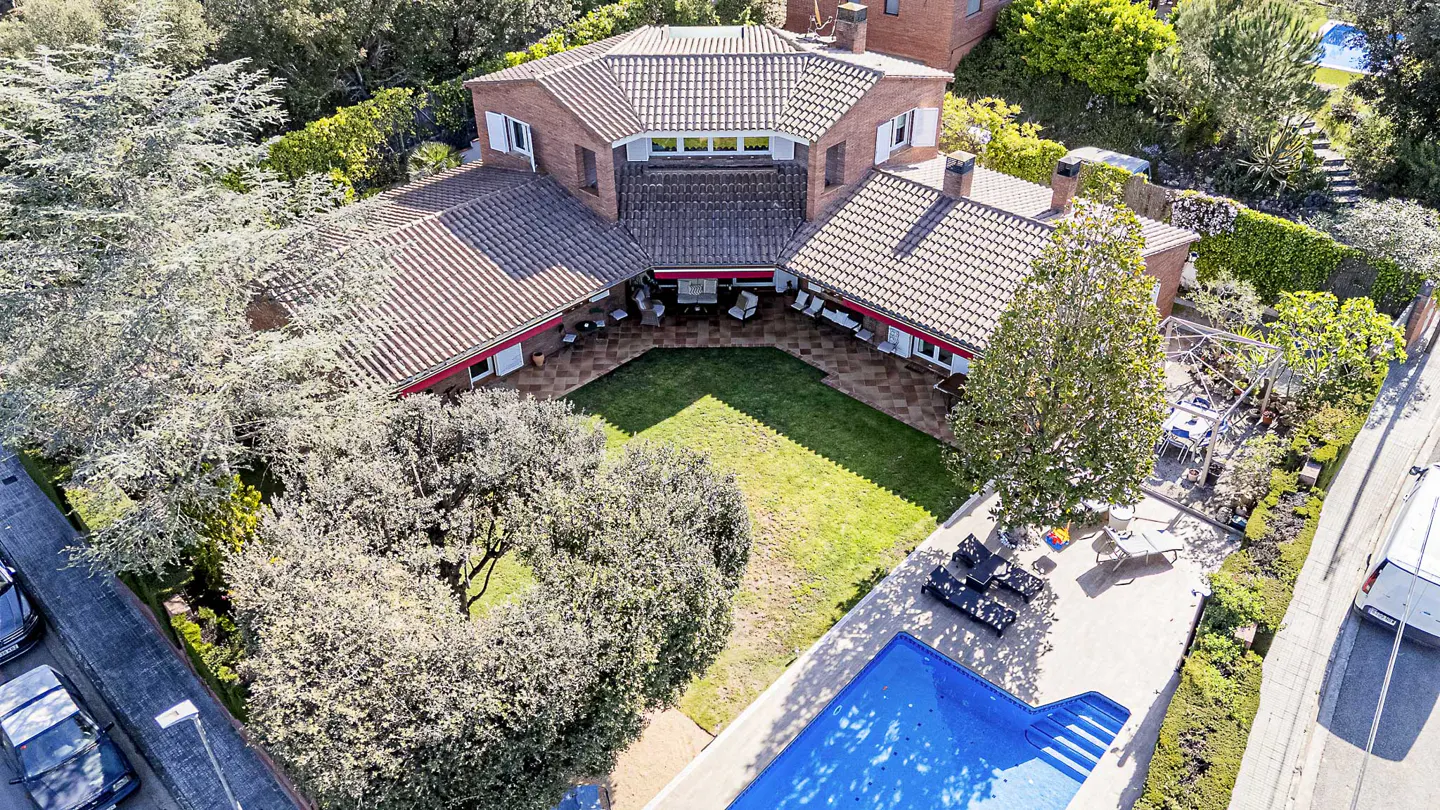 Aerial view of a large brick house with a red tile roof, a pool, and a green lawn surrounded by trees.