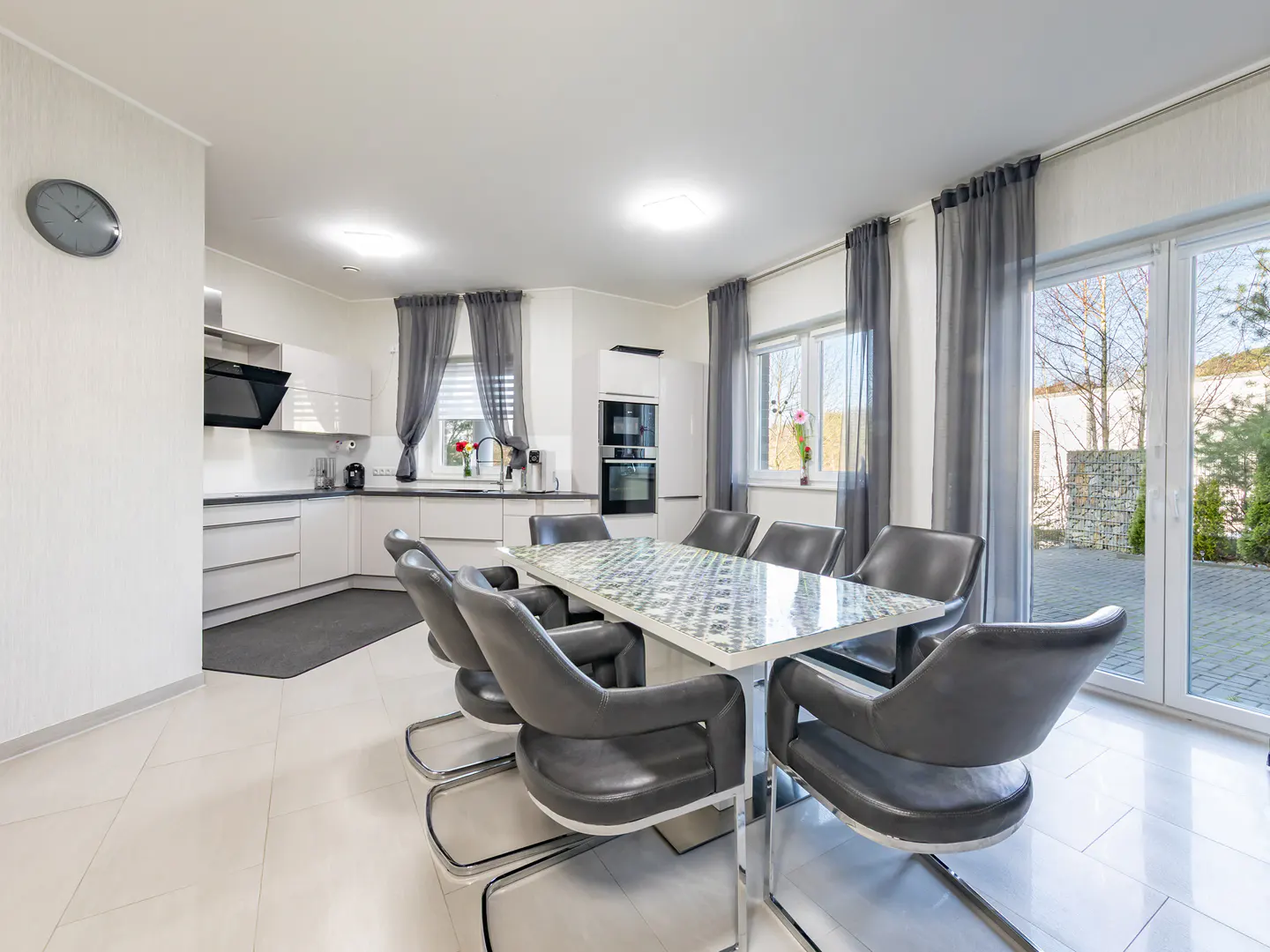 Bright, modern kitchen and dining area with white cabinets, gray chairs, and a glass-topped table. Sliding glass doors lead to a patio.