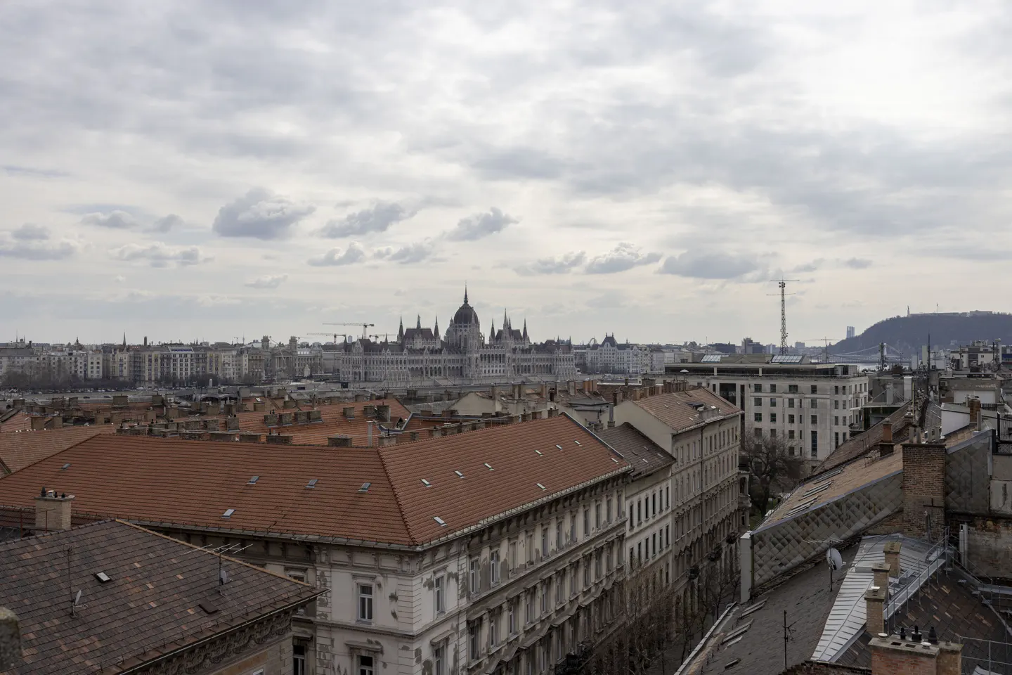 Cityscape view of Budapest, Hungary, featuring red-tiled rooftops and the Hungarian Parliament Building under a cloudy sky.