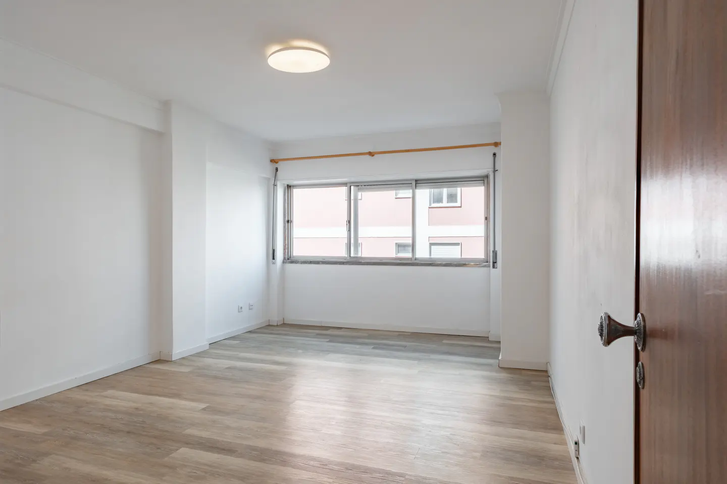 Bright, empty room with light wood floors, white walls, and a large window. A partially open wooden door is on the right.