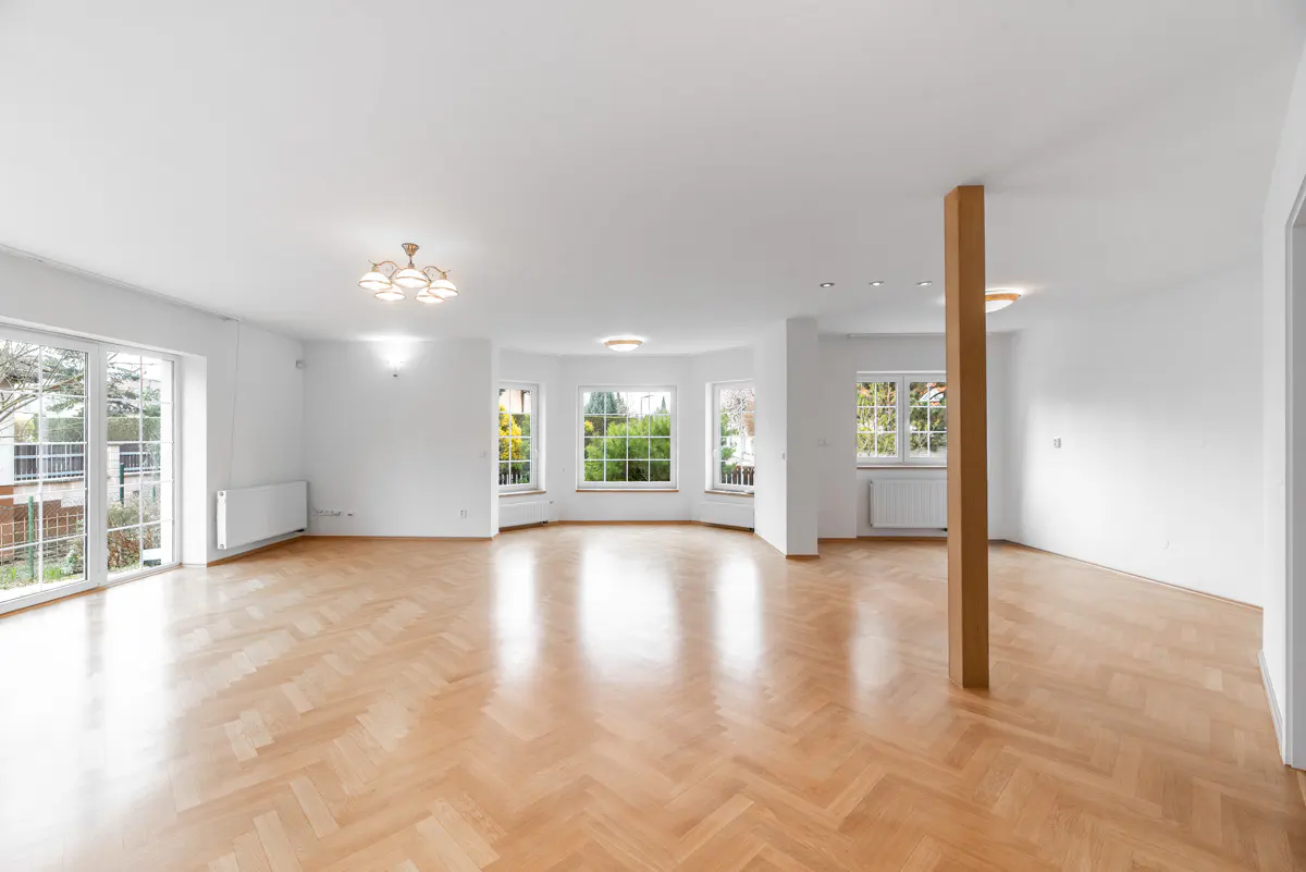 Bright, empty room with herringbone wood floors, white walls, and large windows. A wooden support beam stands near the center.