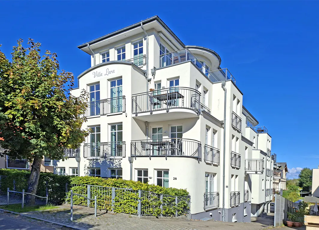 Exterior of "Villa Lara," a multi-story white building with balconies, under a clear blue sky. A green tree is on the left.