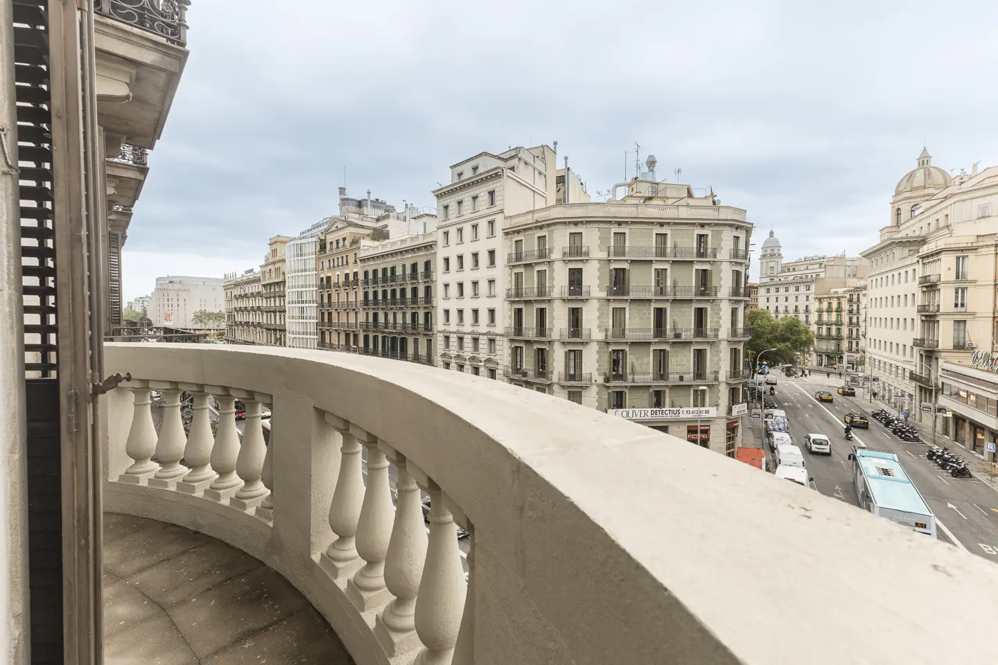 View from a balcony with a stone balustrade overlooking a city street with buildings and traffic.