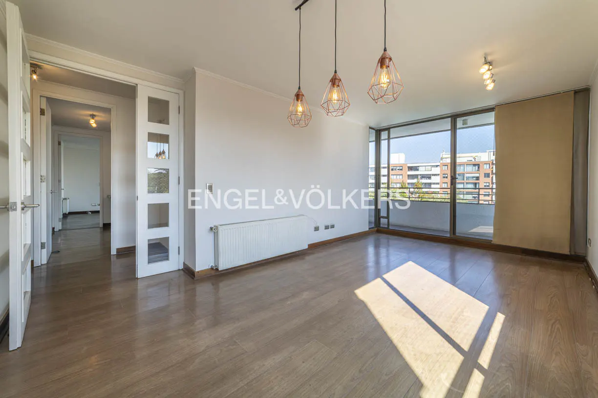 Bright, empty room with wood floors, white walls, and three hanging copper pendant lights. A large window overlooks a balcony and city buildings.