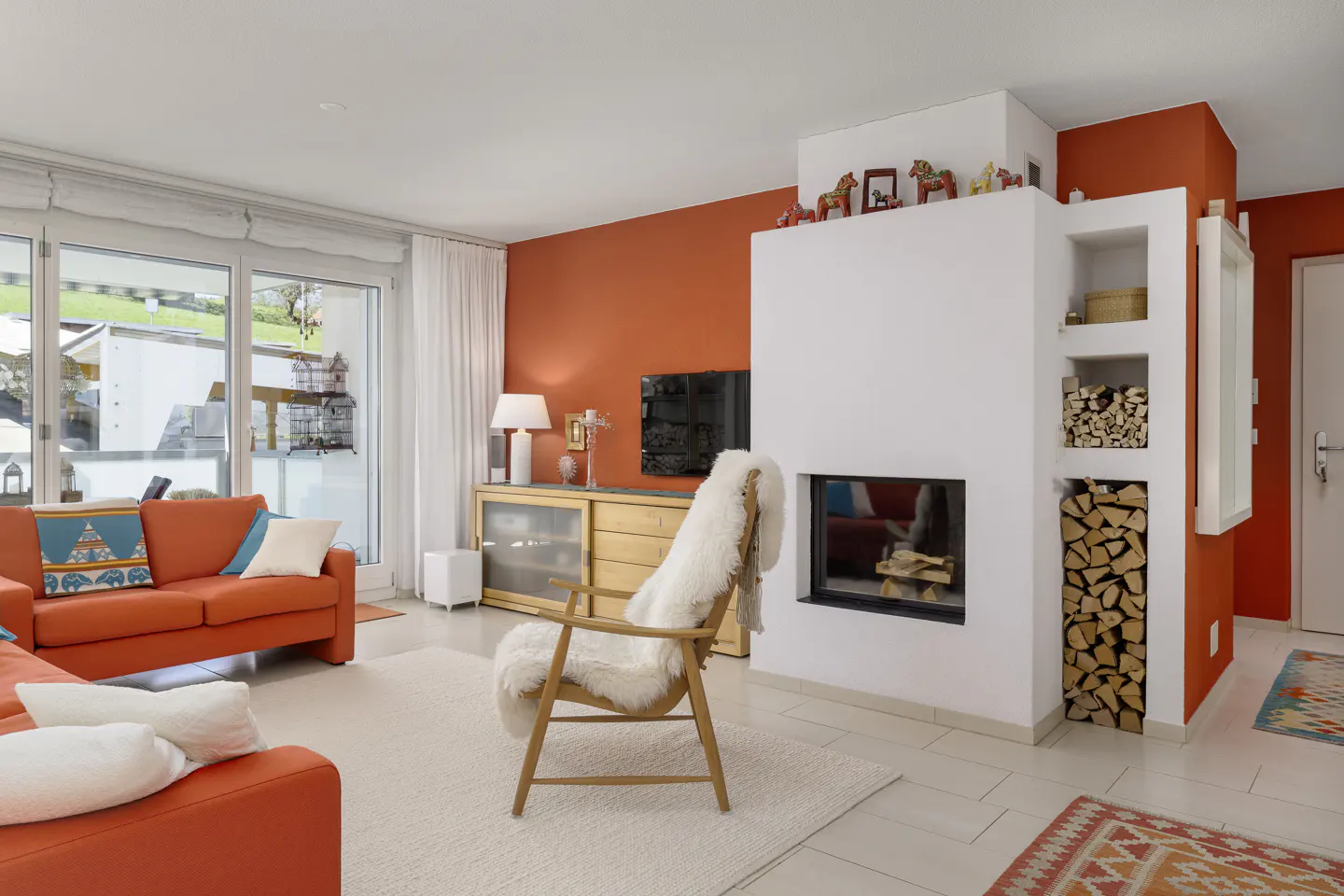 Living room with orange walls, a white fireplace, and a wooden chair with a sheepskin throw. Orange sofa and a sliding glass door to the outside.