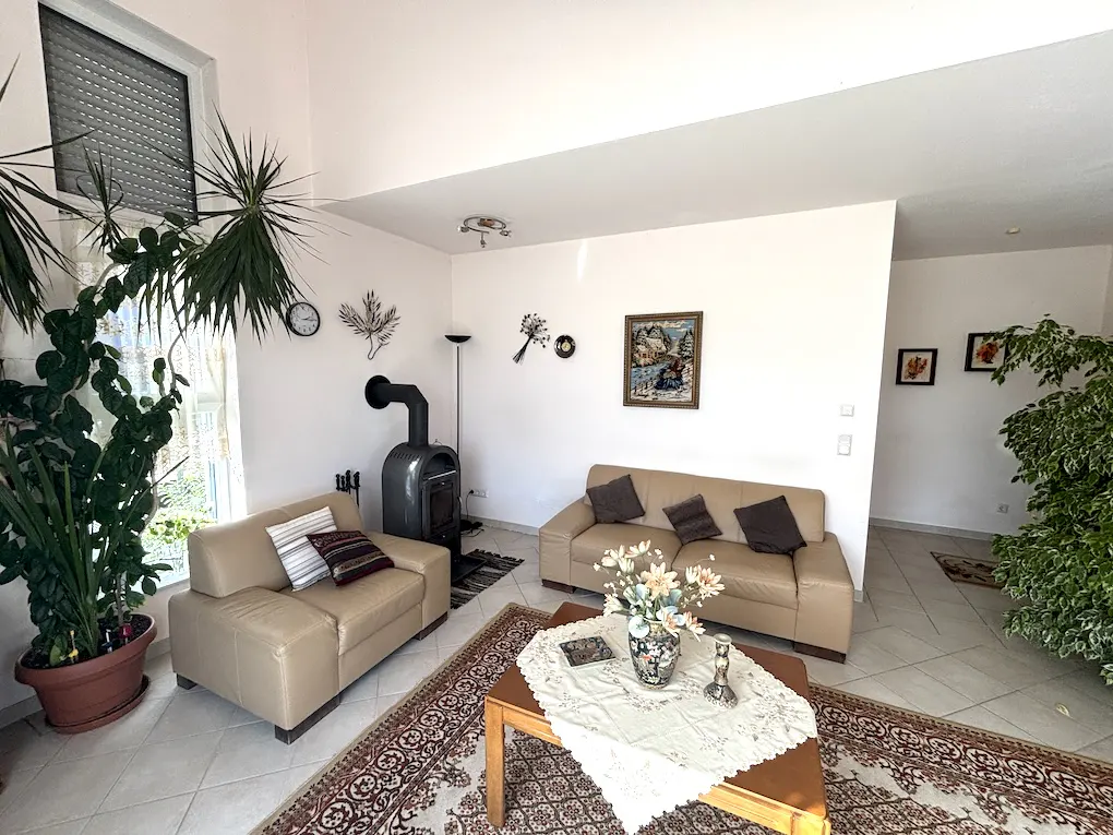 Living room with beige sofas, wood-burning stove, and a brown patterned rug. Plants are near the window.