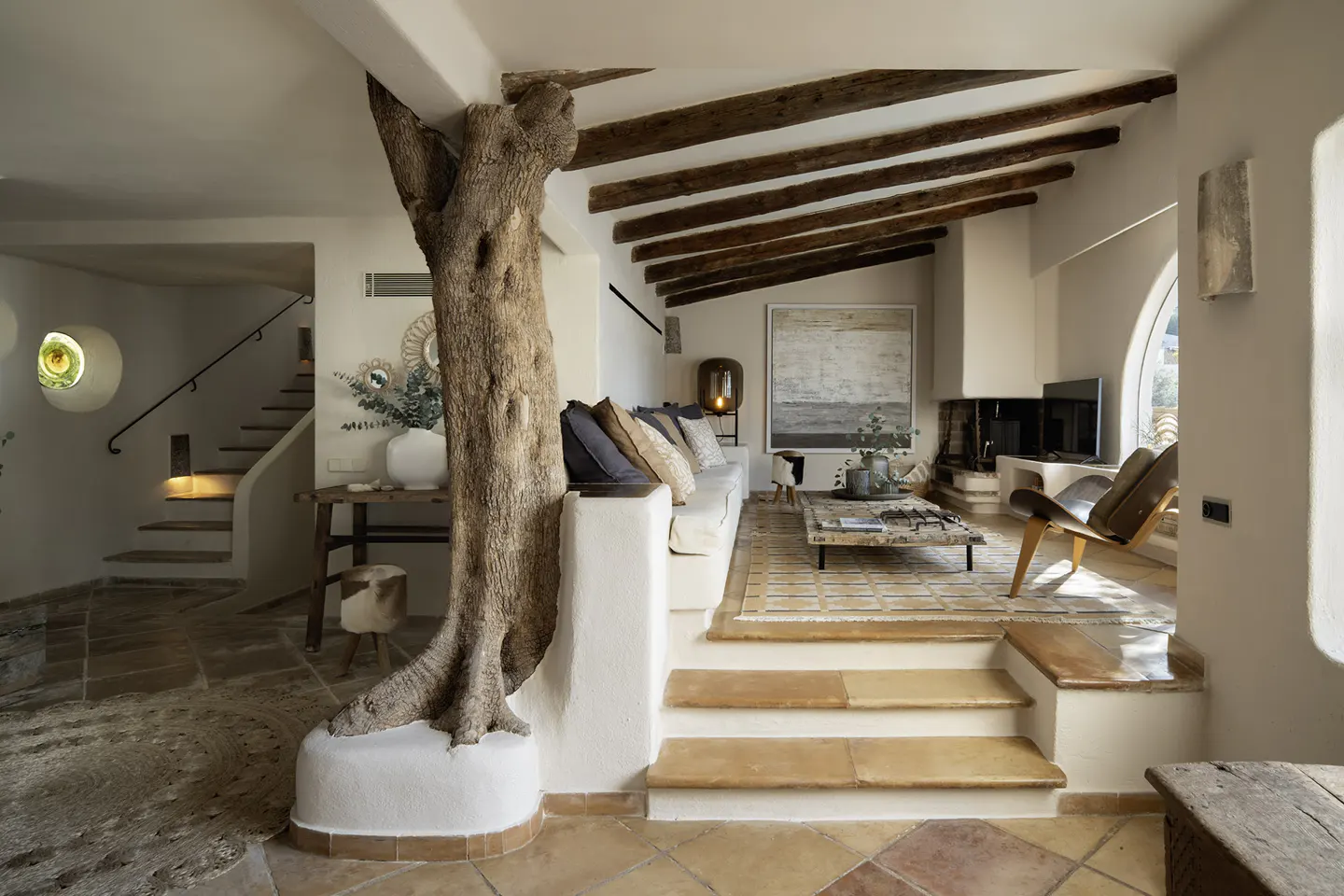 Living room with a tree trunk as a pillar, white sofa, wooden beams, and terracotta tile floor. Stairs lead to another level.