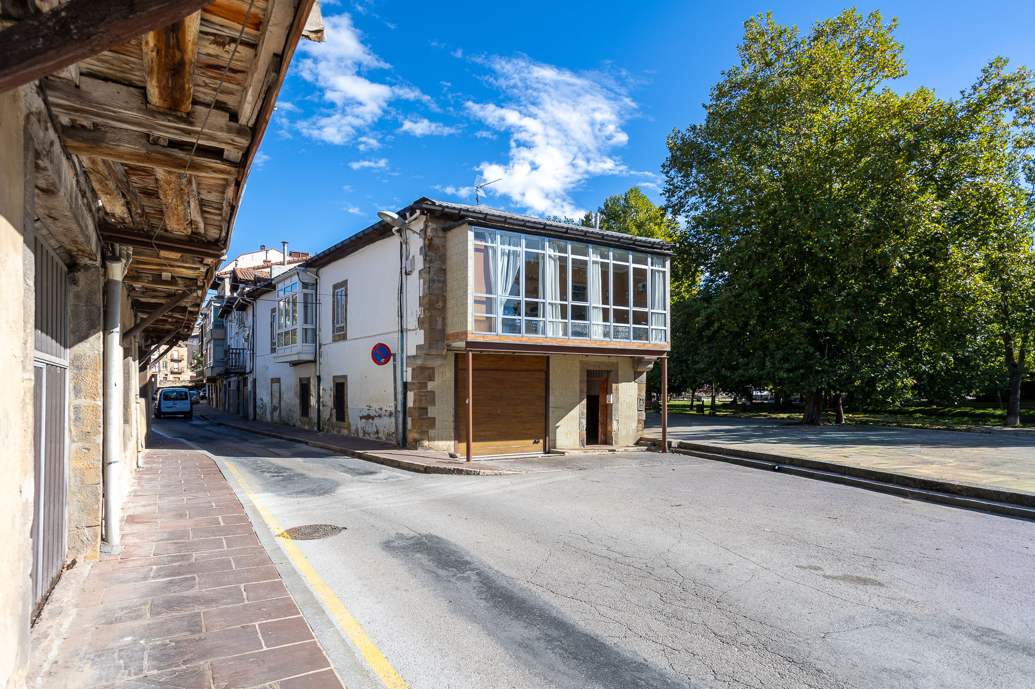 A street view of a white building with a glass-enclosed balcony, a wooden garage door, and a large tree under a blue sky. [AI-generated text]