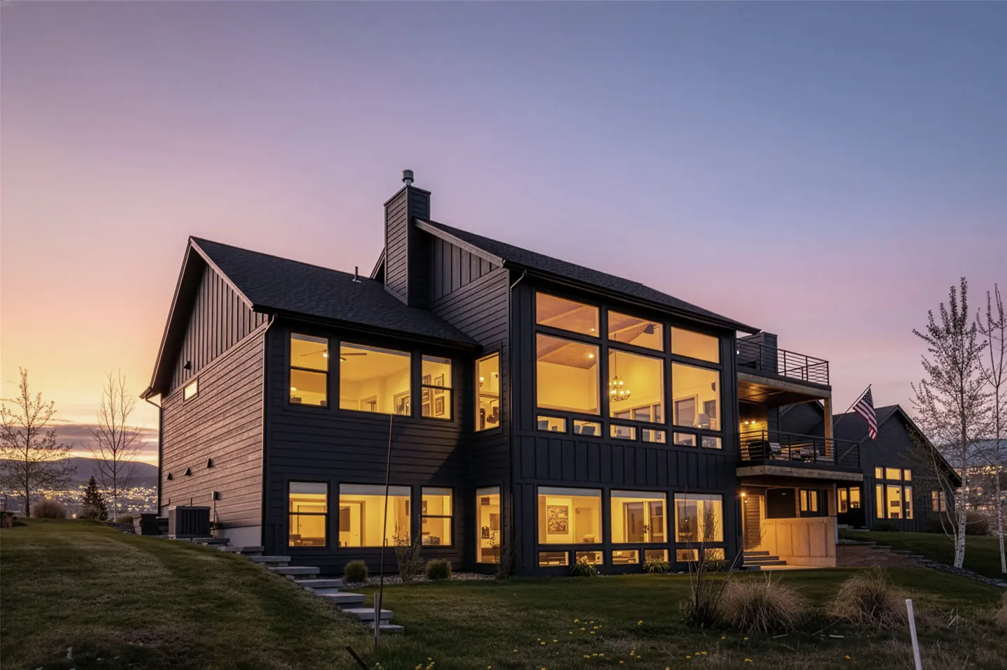 Exterior view of a modern, dark-colored house with large windows at dusk. A green lawn slopes gently towards the house.