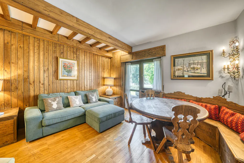 Rustic living room with wood-paneled walls, green sectional sofa, round wooden dining table, and bench seating.