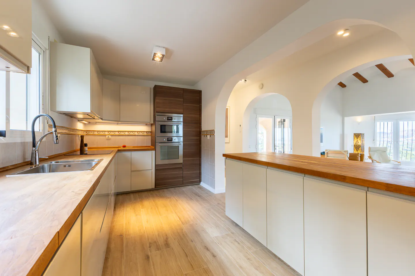 Bright kitchen with white cabinets, wood countertops, stainless steel sink and appliances, and arched doorways leading to a living area.