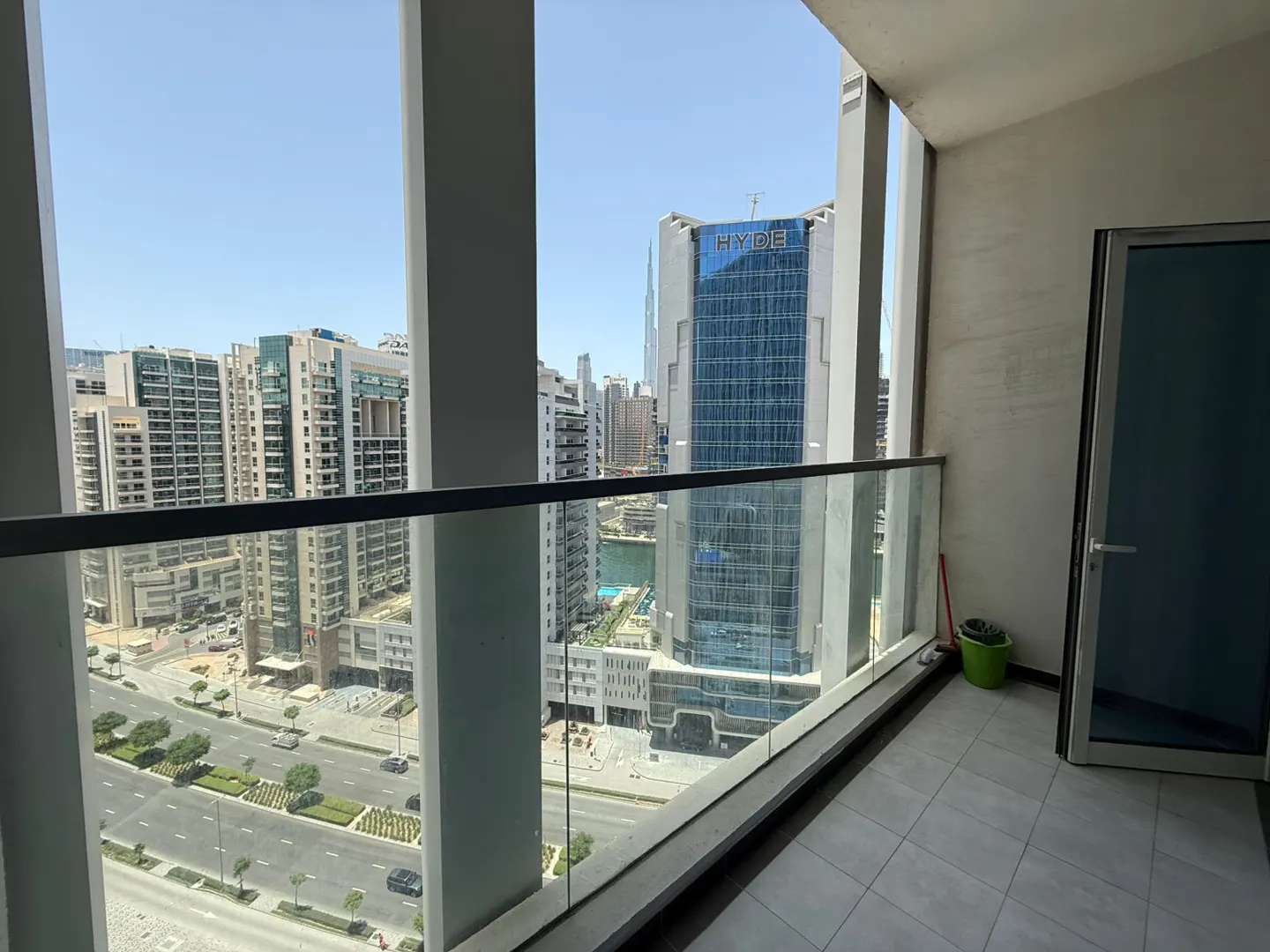 Balcony view of a city with high-rise buildings. Glass railing, gray tile floor, and a blue door are visible.