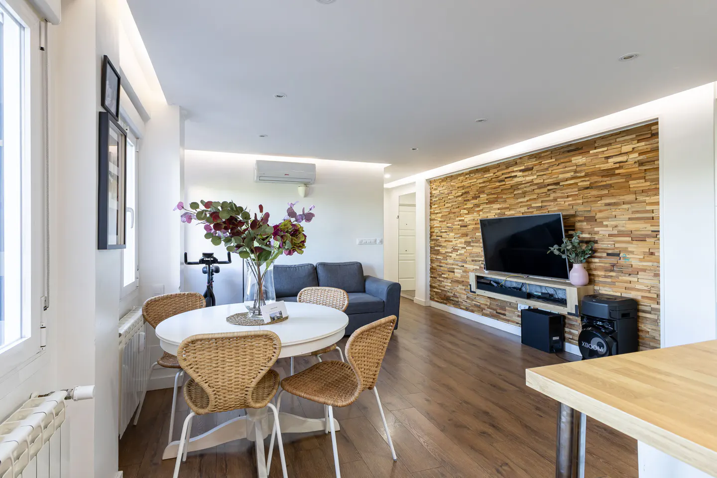 Bright living room with wood floors, a white table with wicker chairs, and a wood-paneled wall with a TV.