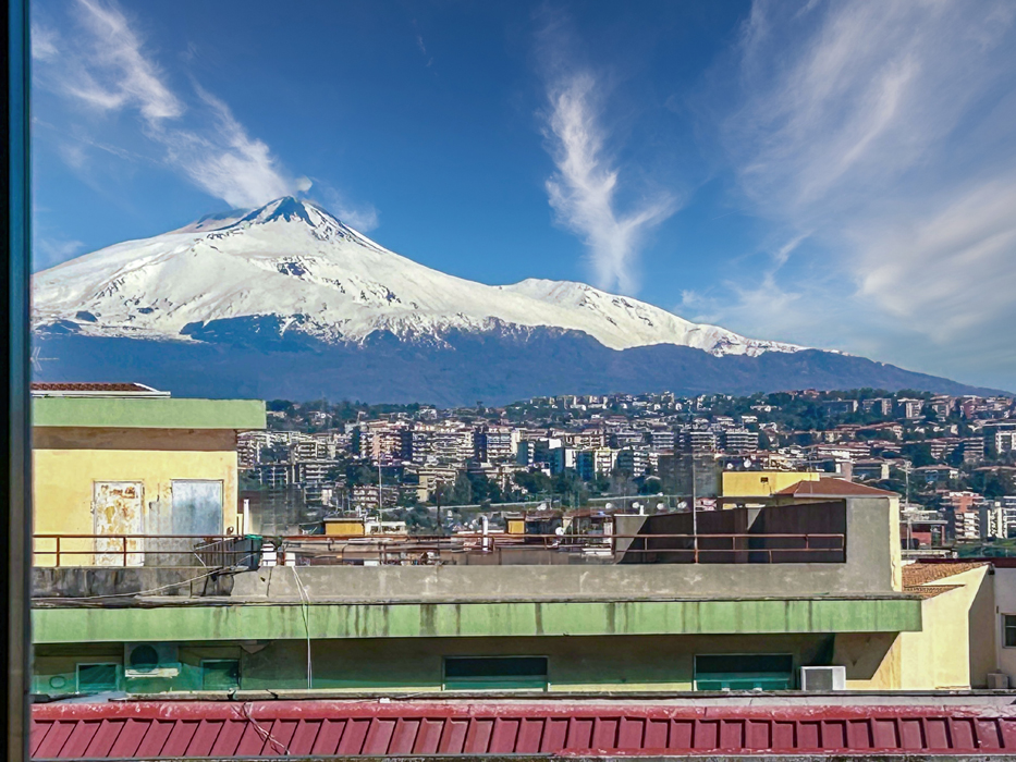 View from a window of Mount Etna, a snow-capped volcano, over rooftops and a city under a blue sky.