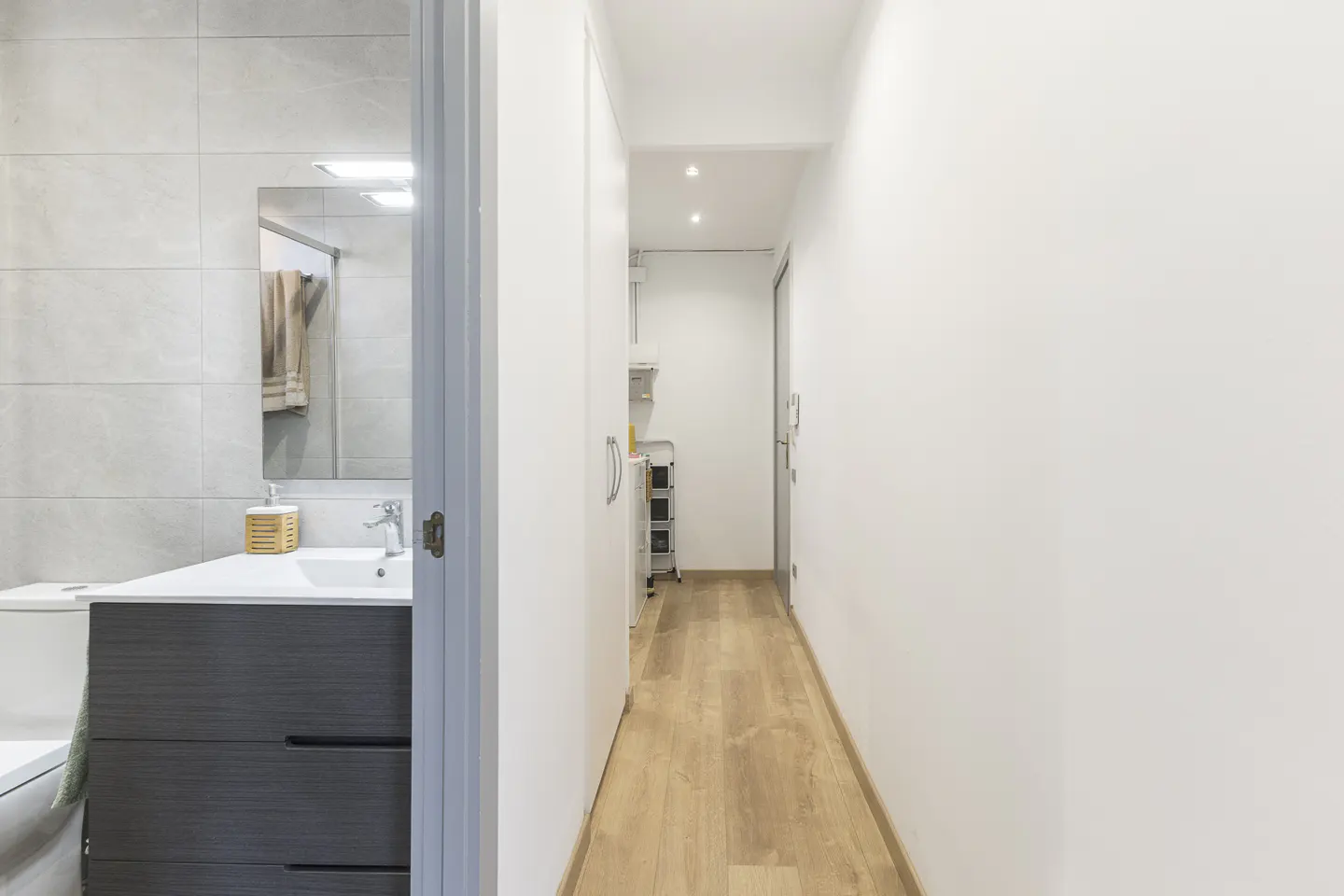 A bright hallway with wood floors leads to a bathroom with gray vanity and tile walls.