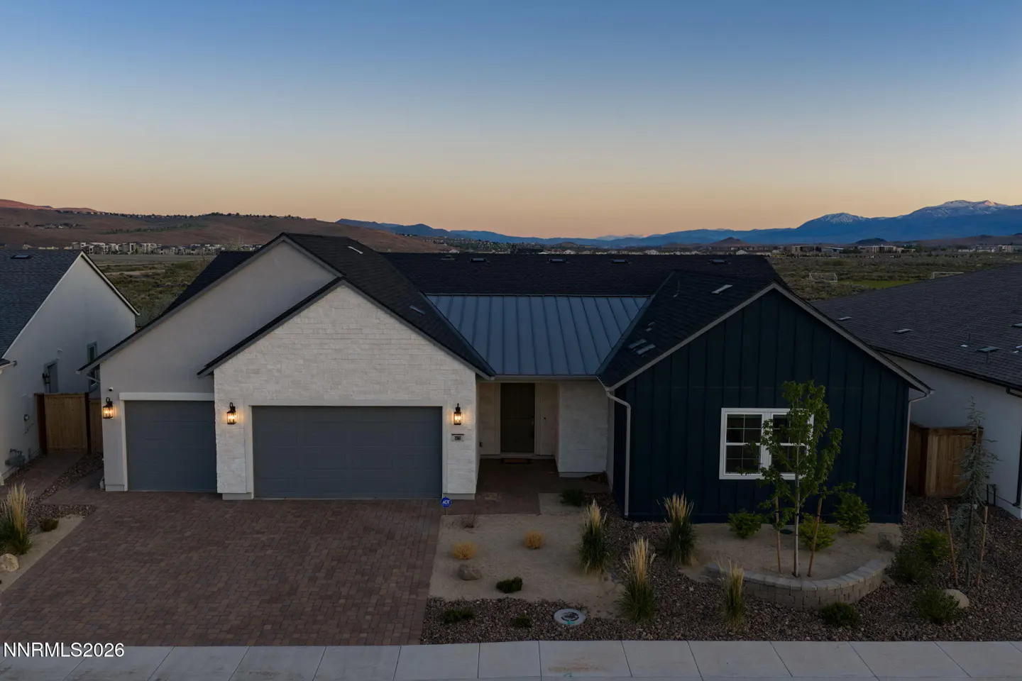 A modern, single-story home with a two-car garage, dark roof, and mixed siding of white and dark blue, set against a mountain backdrop.