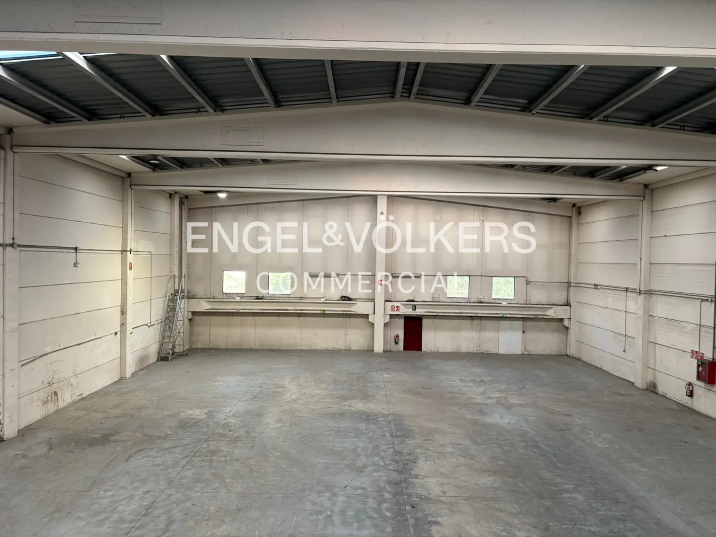 Wide shot of a large, empty warehouse with concrete floors and walls, metal roof, and Engel & Volkers logo.
