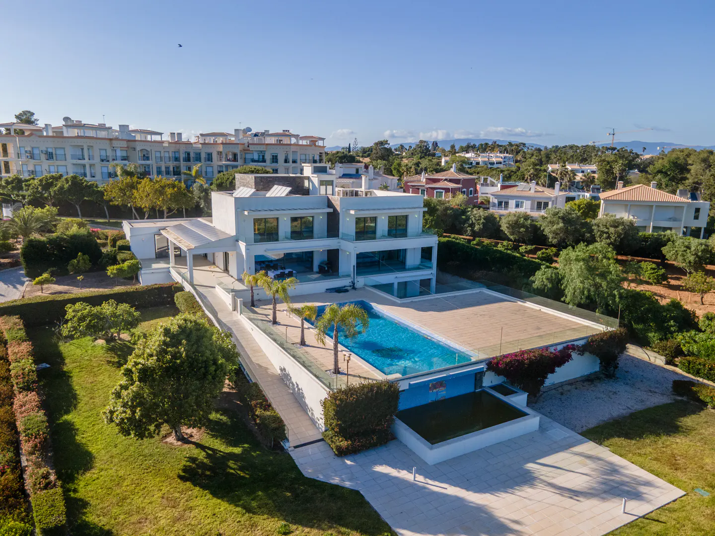 Aerial view of a modern, white two-story house with a blue swimming pool, palm trees, and green lawn. Other houses are visible in the background.