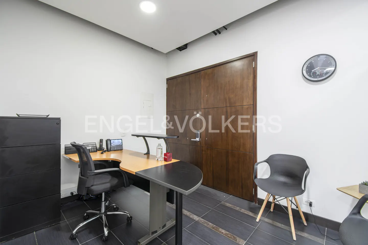 Modern office with a curved desk, black rolling chair, dark wood double doors, and a wall clock on a white wall.