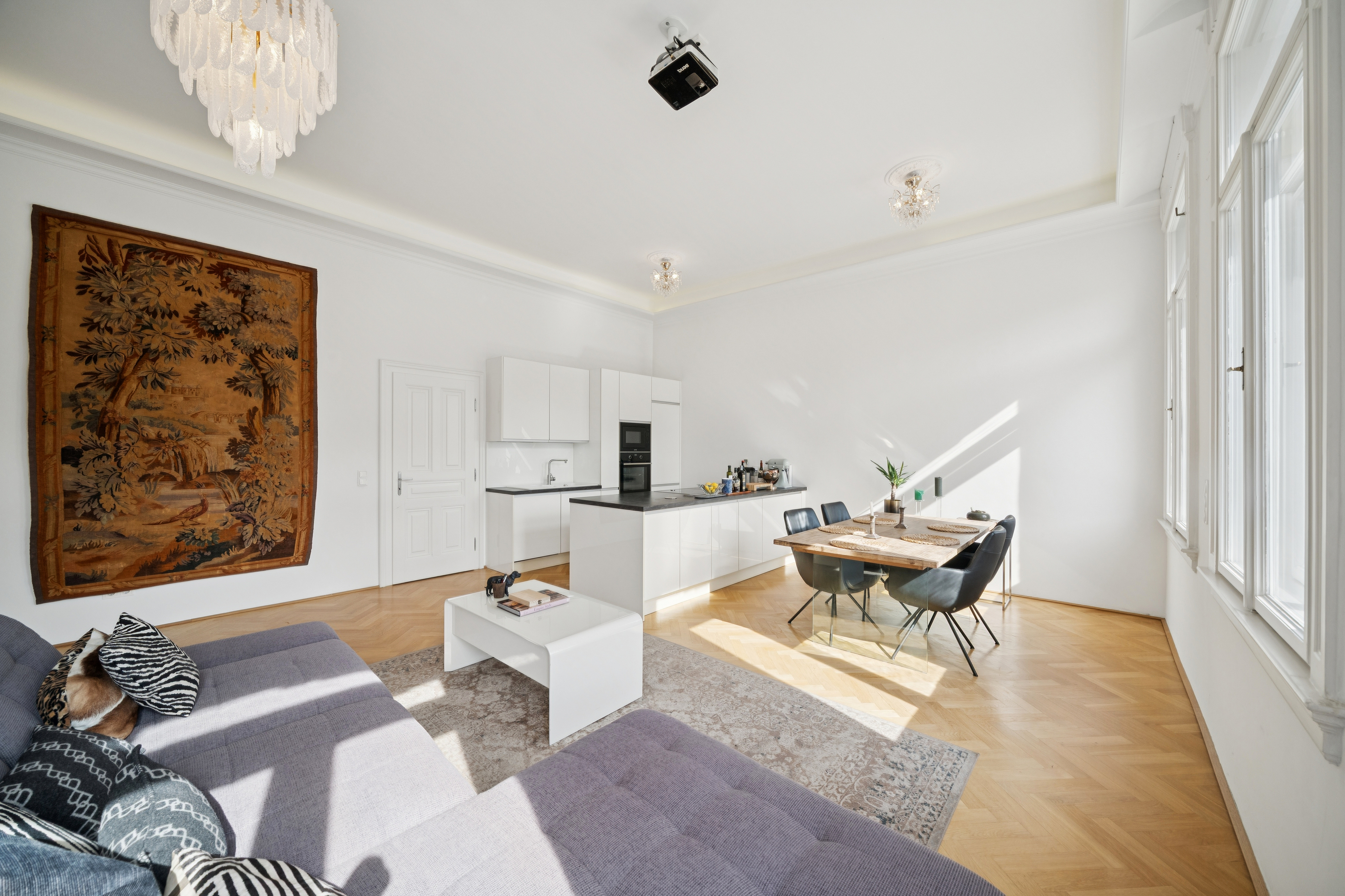 Bright open-plan living area with a grey sectional sofa, white kitchen, dining table, and a large wall tapestry. [AI-generated text]