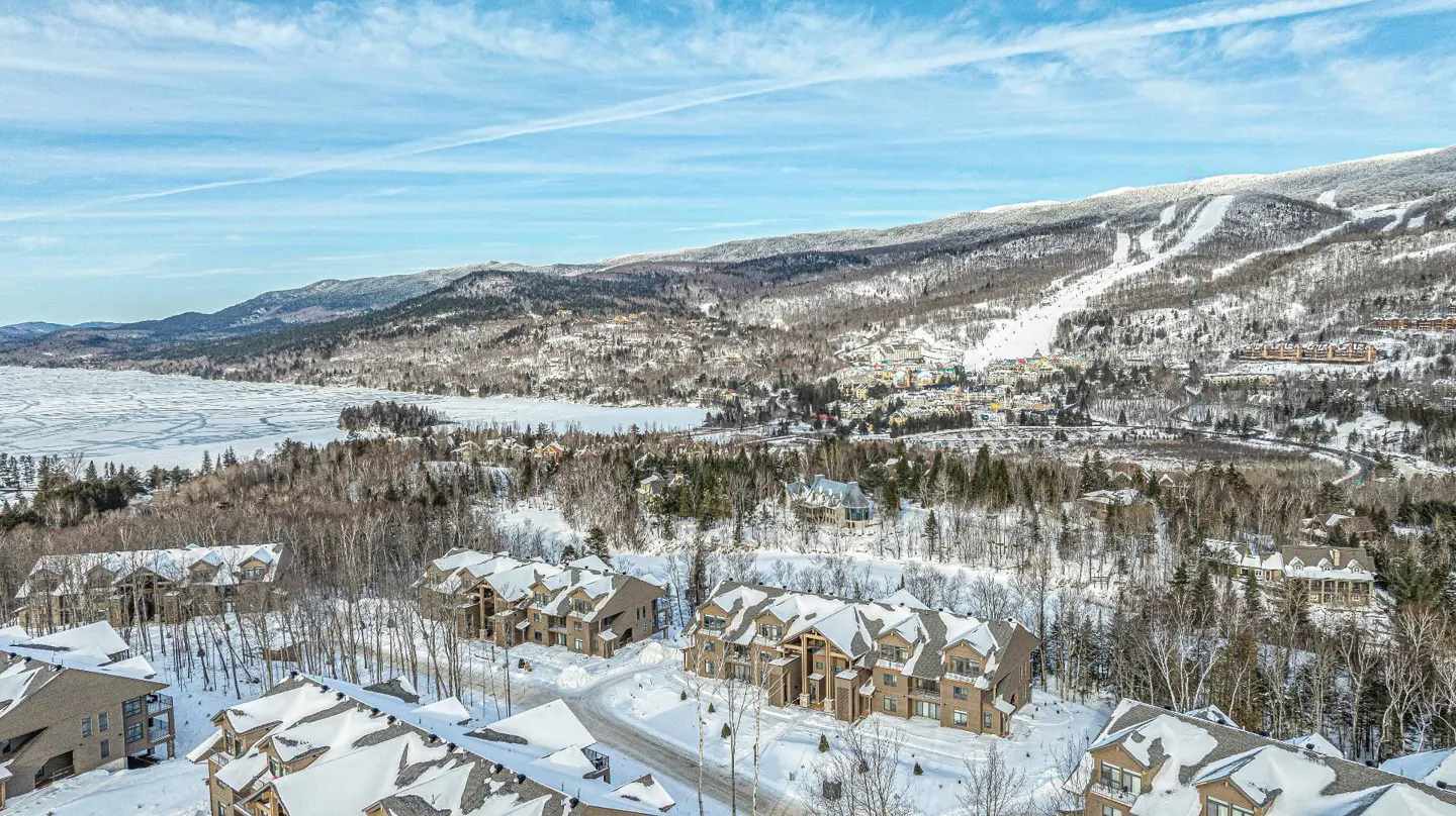 Aerial view of brown condos with snow-covered roofs, a frozen lake, and a ski mountain under a blue sky.