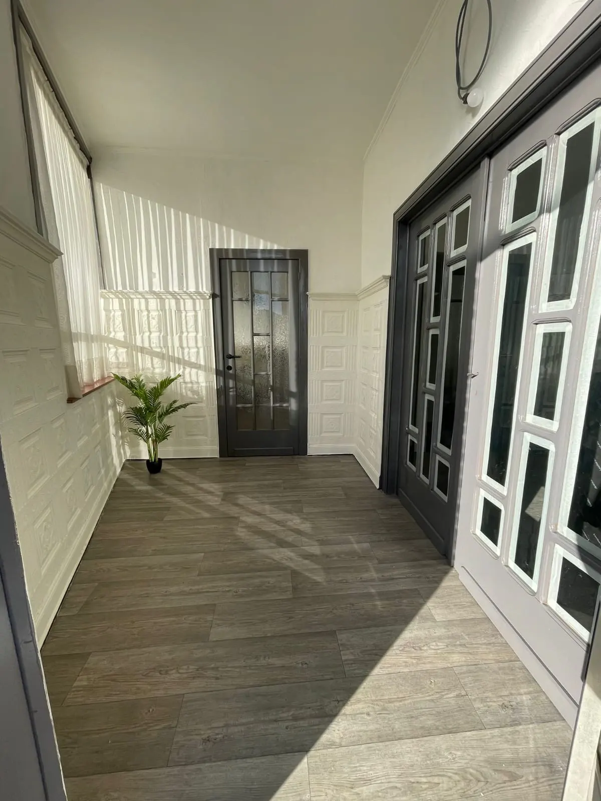 A bright foyer with gray wood-look floors, white walls, and two gray doors. A small potted plant sits near a window with sheer white curtains.