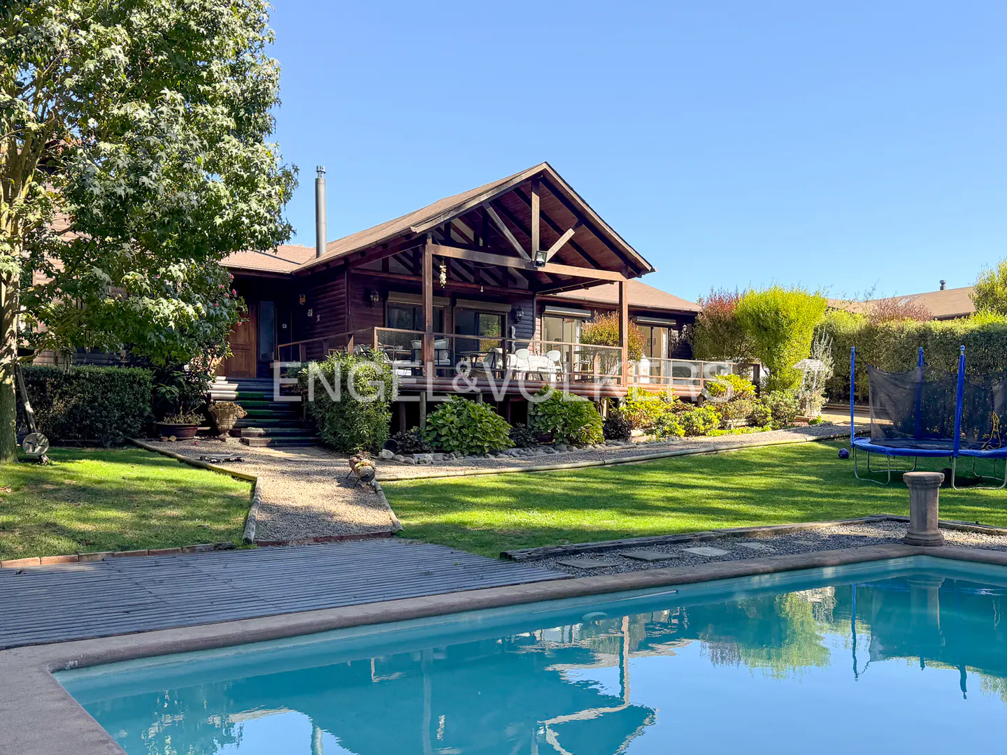 Exterior shot of a brown wooden house with a pool in the foreground and a green lawn.