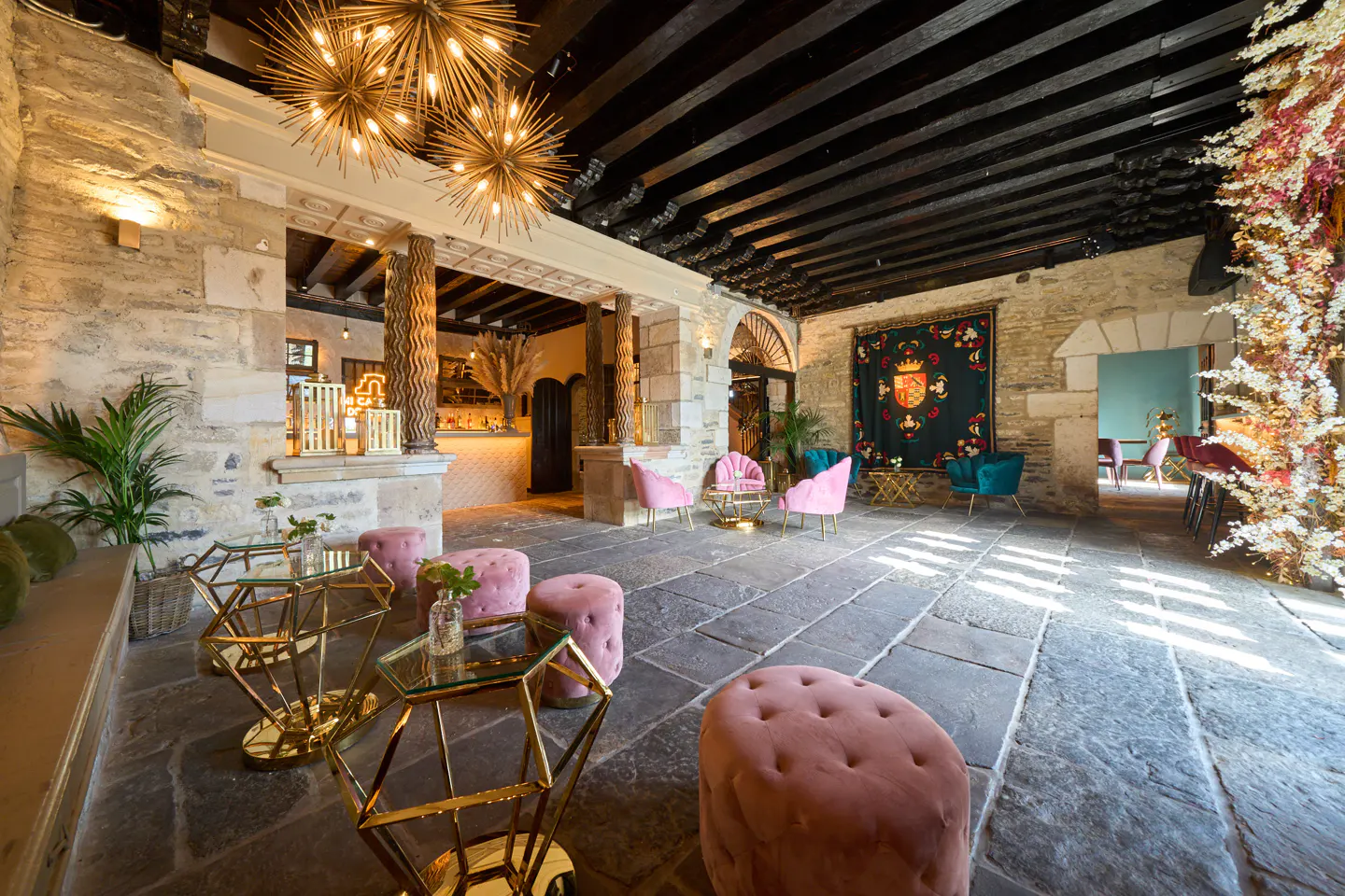 Interior view of a stone-walled room with pink seating, gold tables, and a dark wood beamed ceiling.