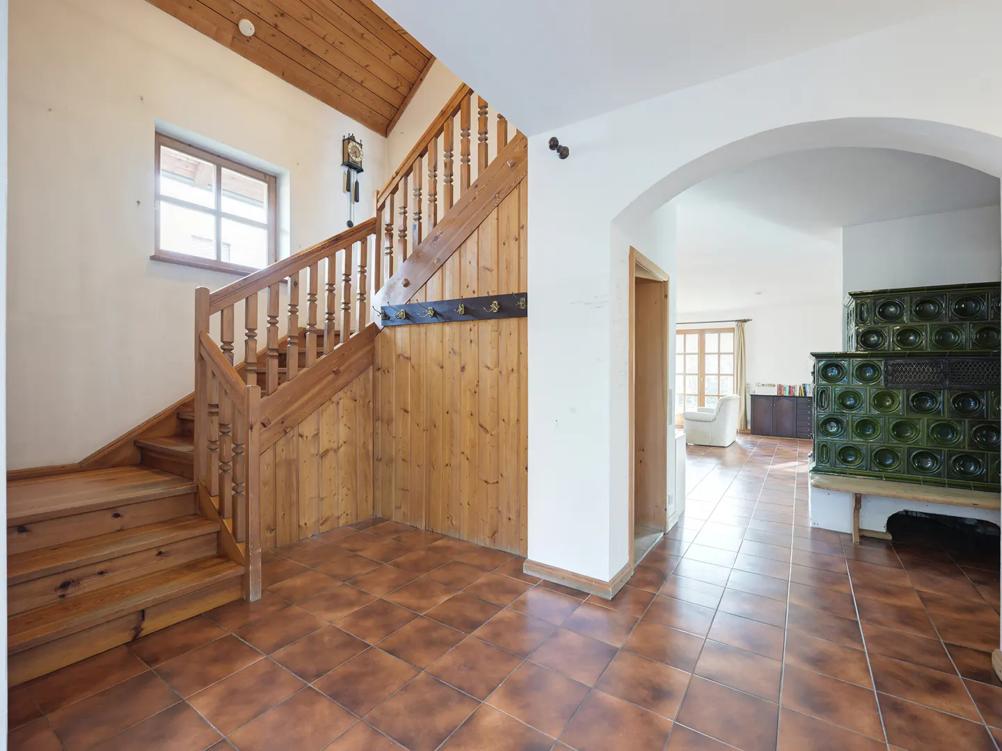 Interior view of a home's foyer with a wooden staircase, brown tiled floor, and a green tiled stove.