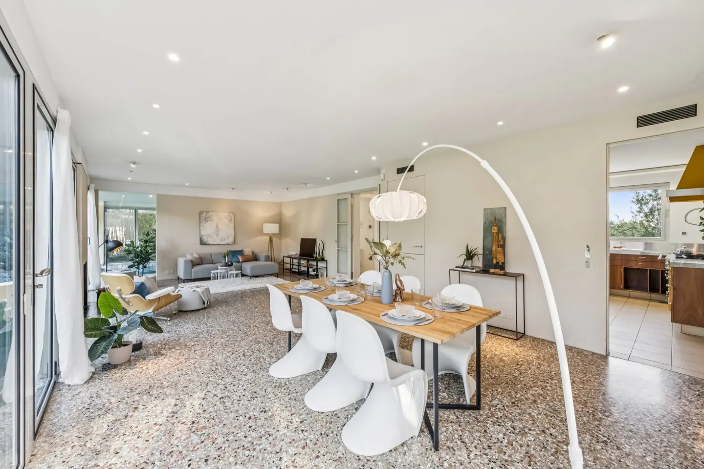 Bright, open-plan living and dining area with terrazzo flooring. A wooden table with white chairs sits under an arc lamp. A grey sofa is visible in the background.