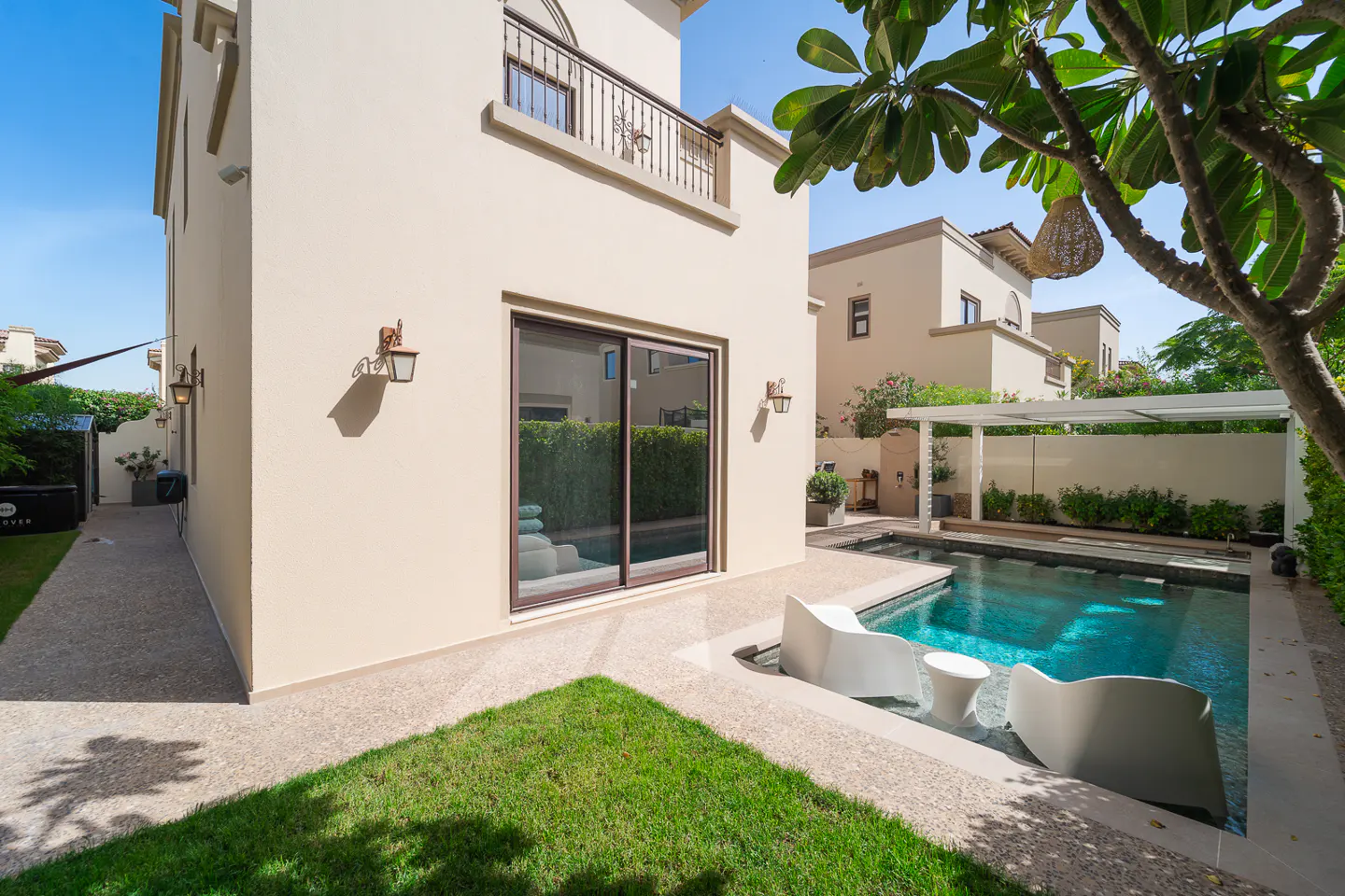 Backyard view of a two-story beige house with a pool, white chairs, and green grass.