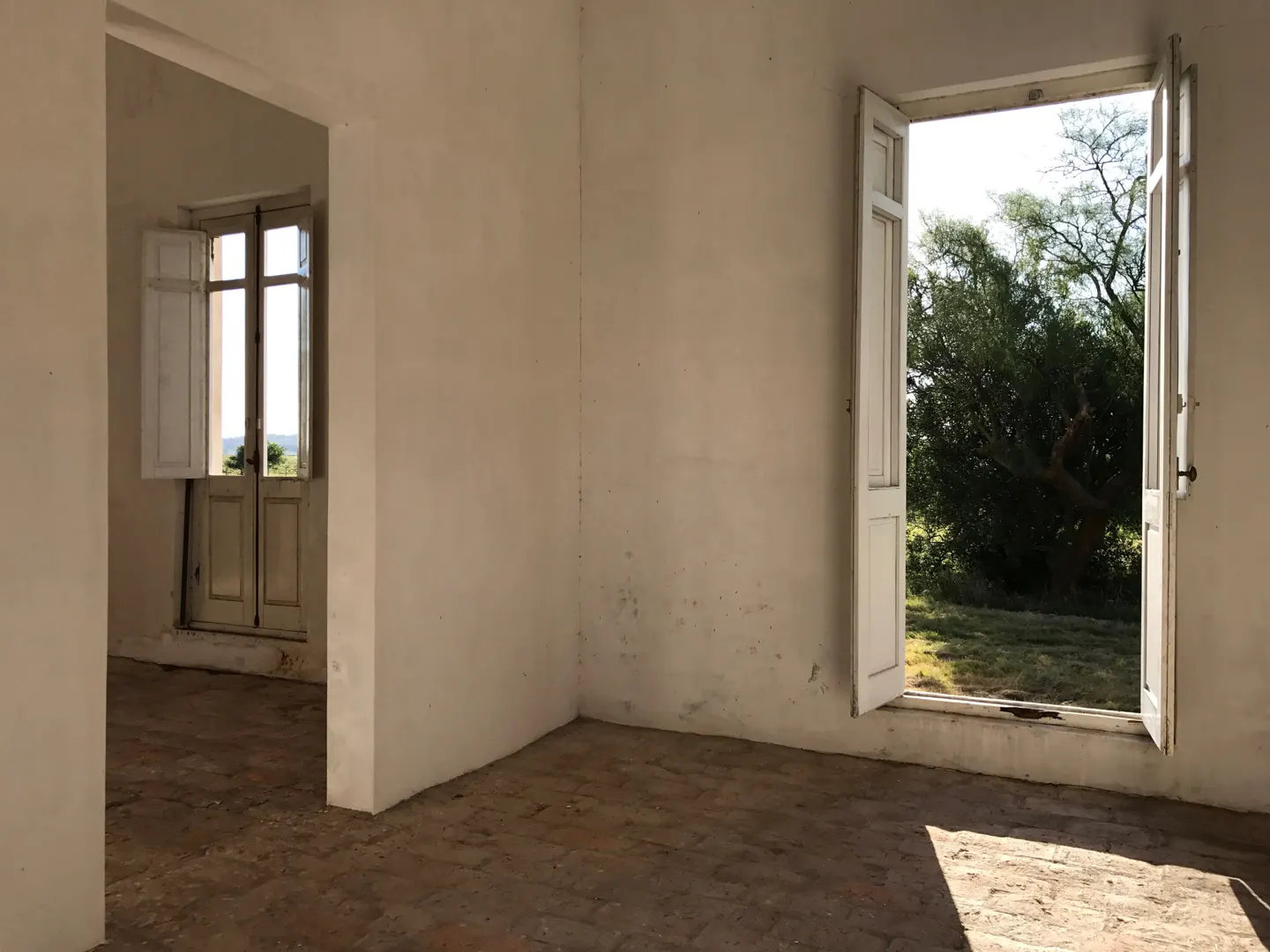 Empty room with white walls, terracotta tile floors, and open white doors looking out onto a sunny, tree-filled landscape.