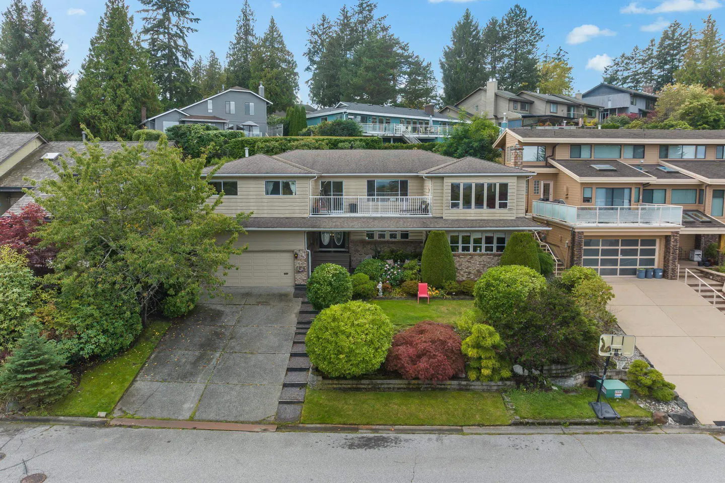 Two-story beige house with a balcony, garage, and landscaped yard. Other houses are visible on a hill in the background.