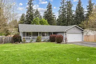 A gray single-story house with a white porch and garage sits on a green lawn with trees in the background.