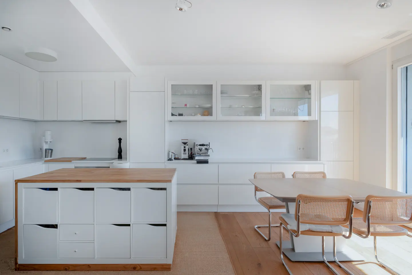 Modern white kitchen with a wooden island, glass-front cabinets, and a dining table with four cane-back chairs.