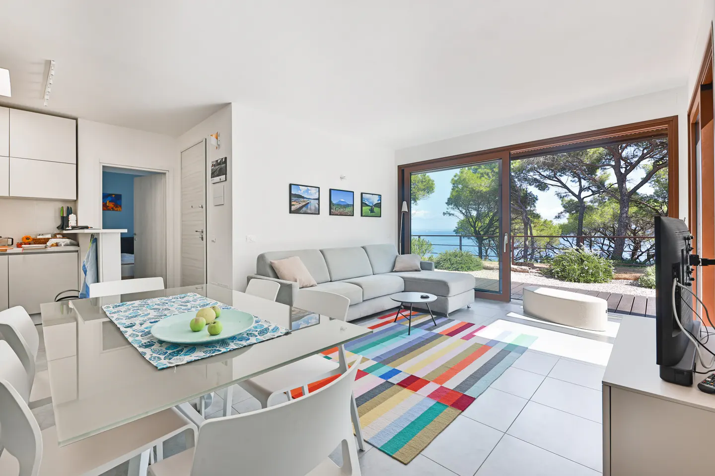 Bright, modern living room with a gray sofa, colorful rug, and a glass dining table. Large windows offer a view of trees and the sea.