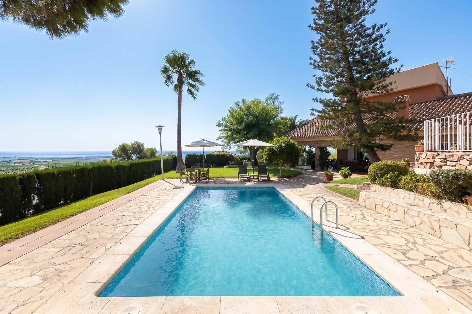 A rectangular swimming pool with stone decking, lounge chairs, and umbrellas, set against a scenic coastal landscape. [AI-generated text]