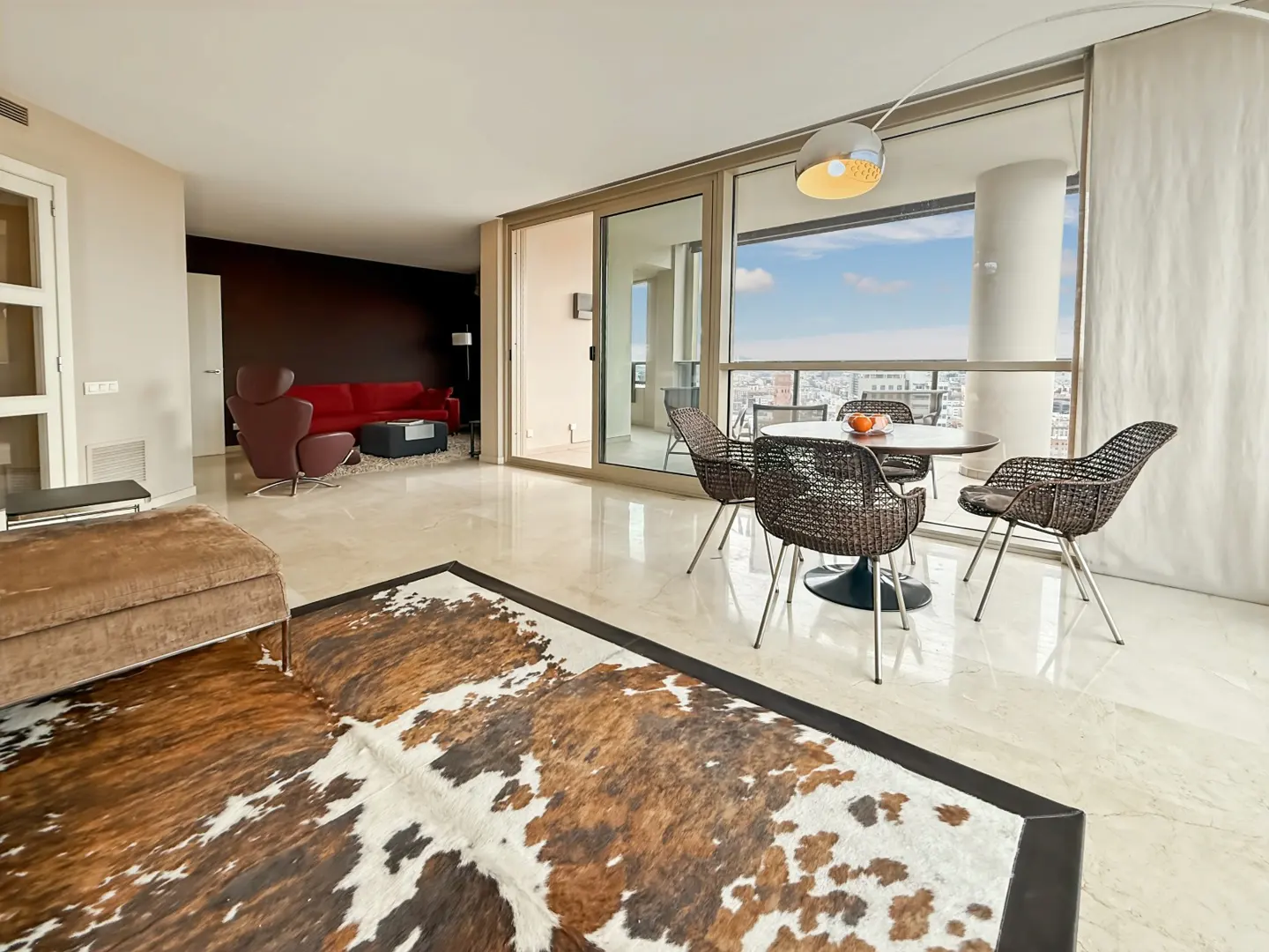 A modern living room with a brown and white cowhide rug, a dining table with four chairs, and a red sofa in the background.