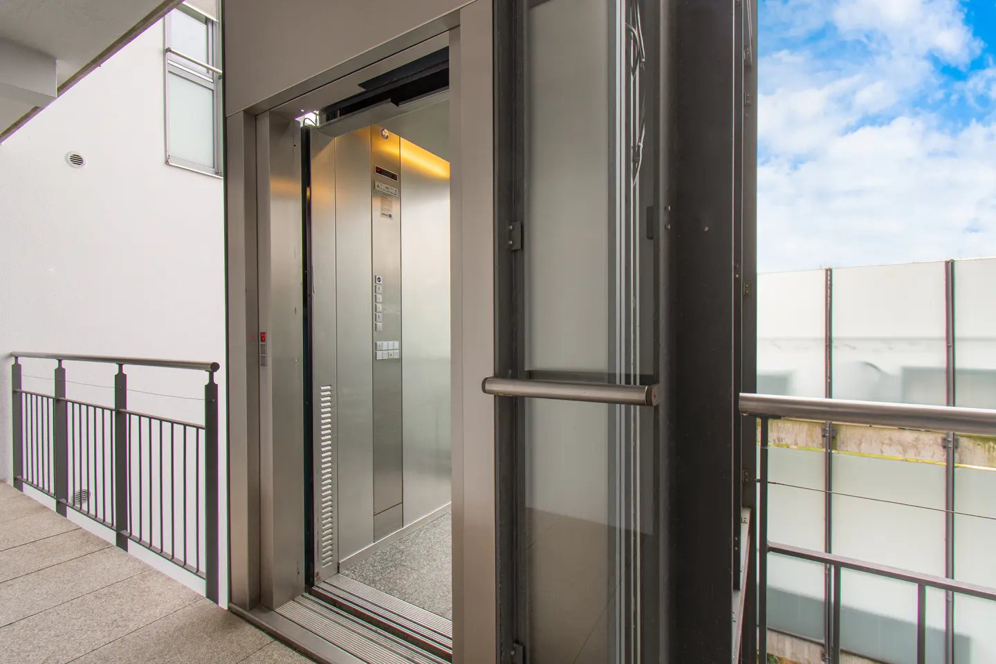 An open elevator with stainless steel interior and exterior, on a balcony with a metal railing, and a blue sky in the background.