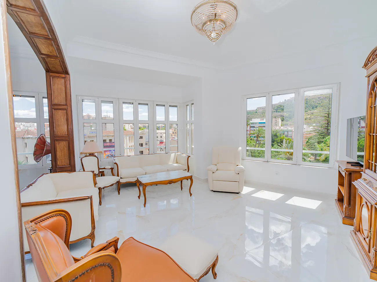 Bright living room with white marble floors, cream furniture, wooden accents, a crystal chandelier, and large windows.