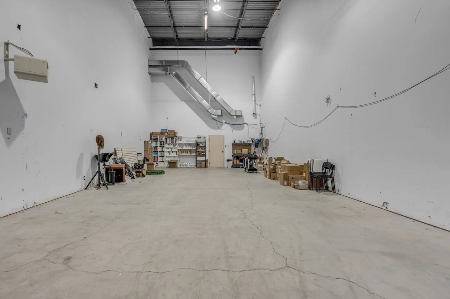 Wide shot of a large, empty commercial space with white walls, concrete floors, and exposed ductwork. Shelves and boxes line the back walls.