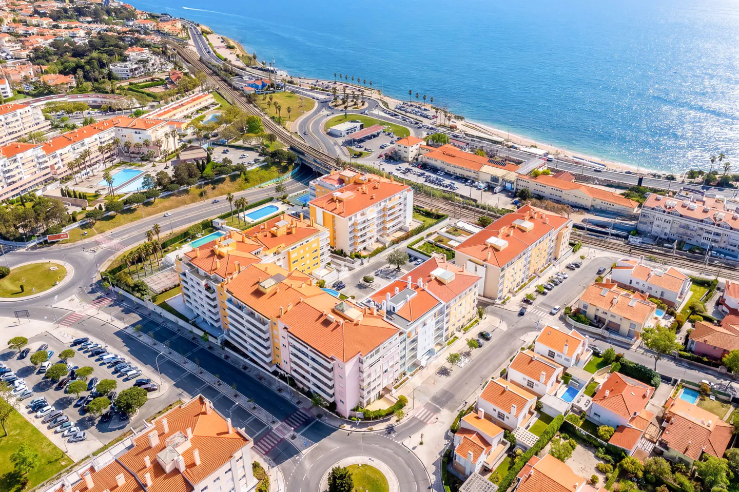 Aerial view of a coastal town with orange-roofed buildings, roundabouts, and a blue ocean backdrop.