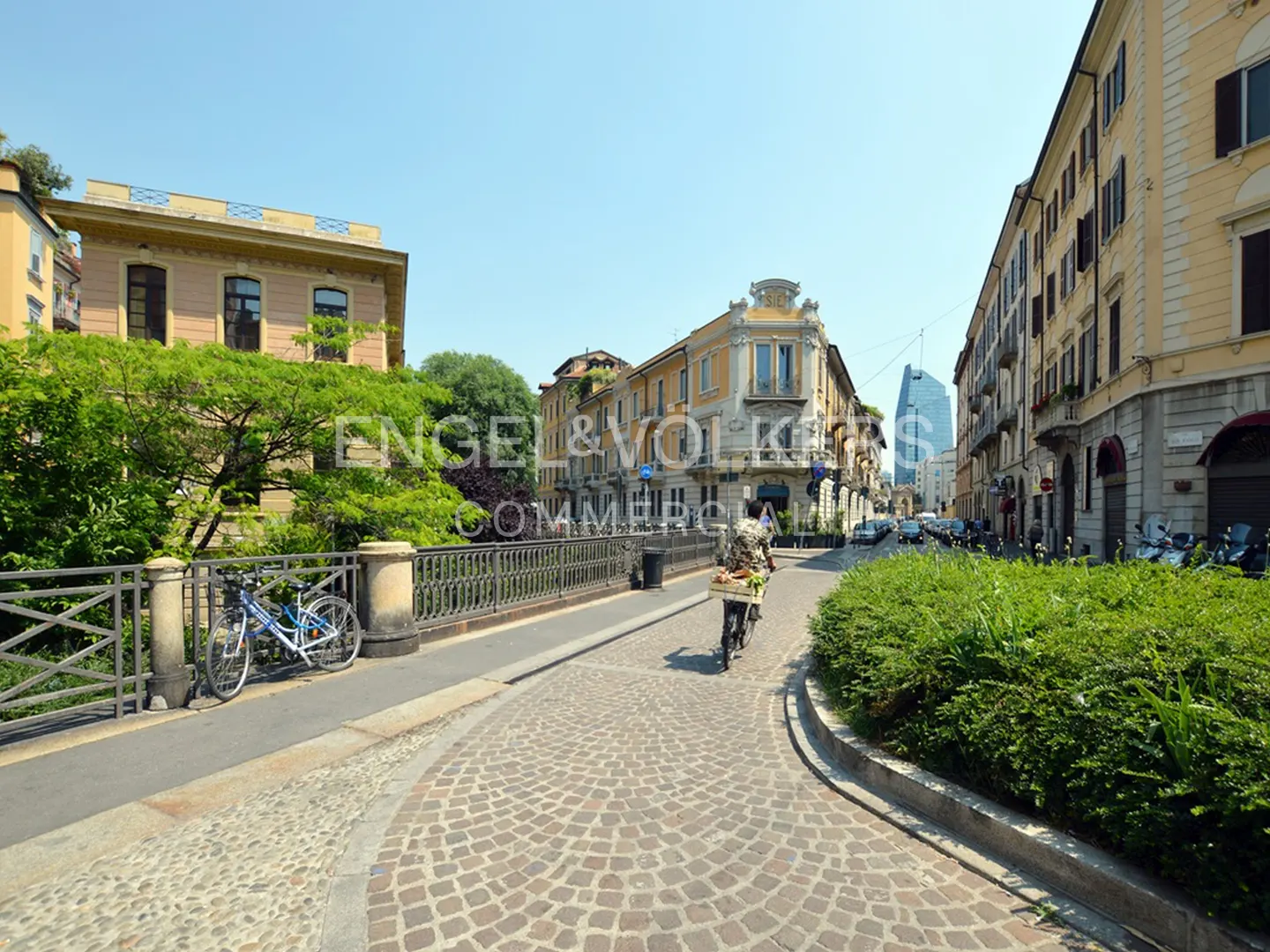 Cobblestone street view in Milan, Italy. A cyclist rides past green bushes and buildings under a clear blue sky.