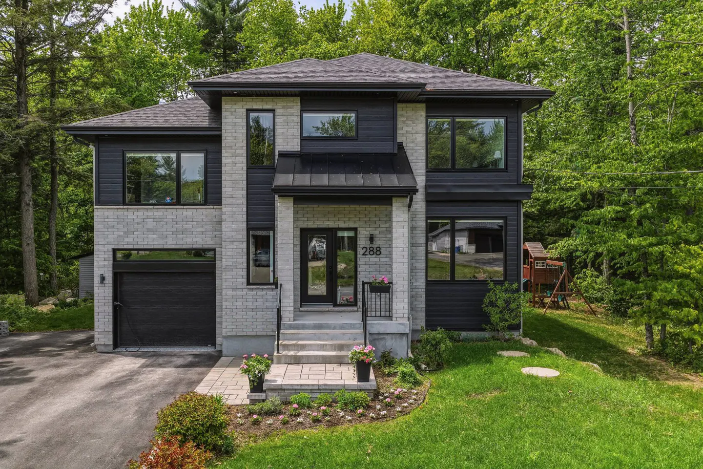 Modern two-story home with light gray brick, black siding, a single garage, and a lush, wooded backyard setting.