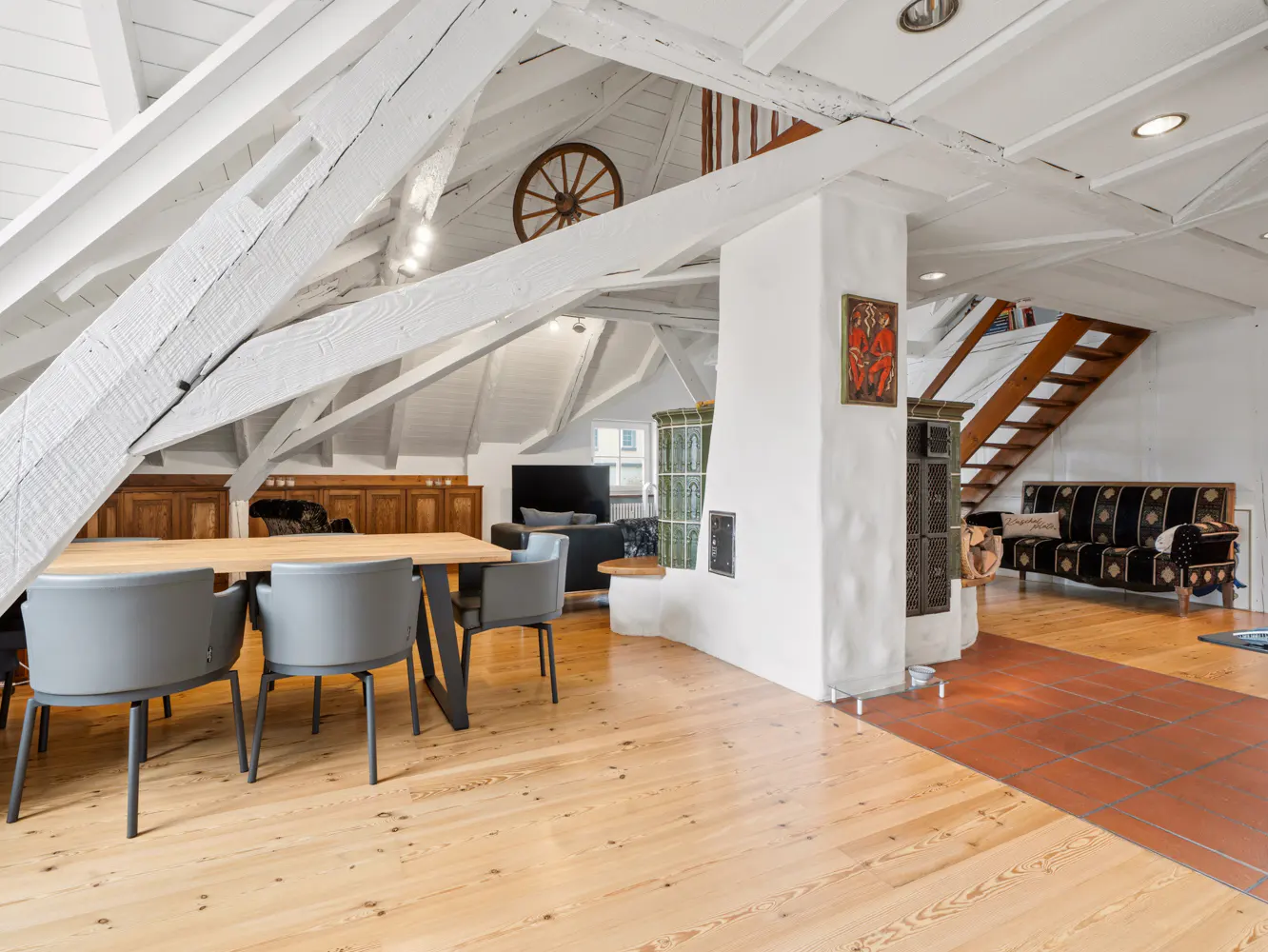 Attic apartment with exposed white beams, wood floors, and a dining table with gray chairs. A decorative wheel hangs on the wall.