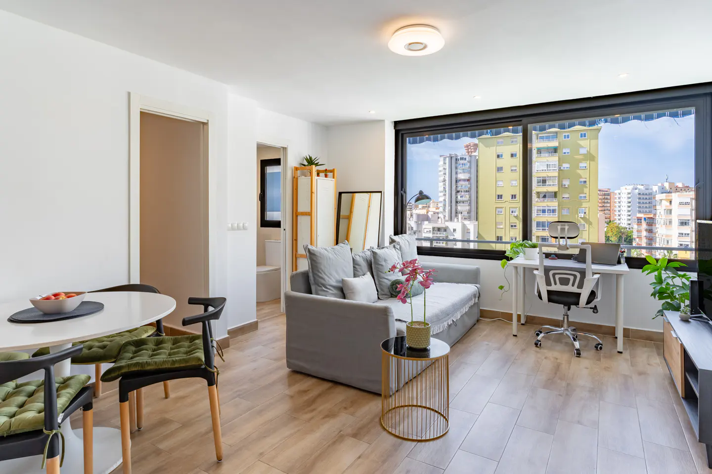 Bright living room with a gray sofa, desk, and city view through a large window. A round table and chairs are on the left.