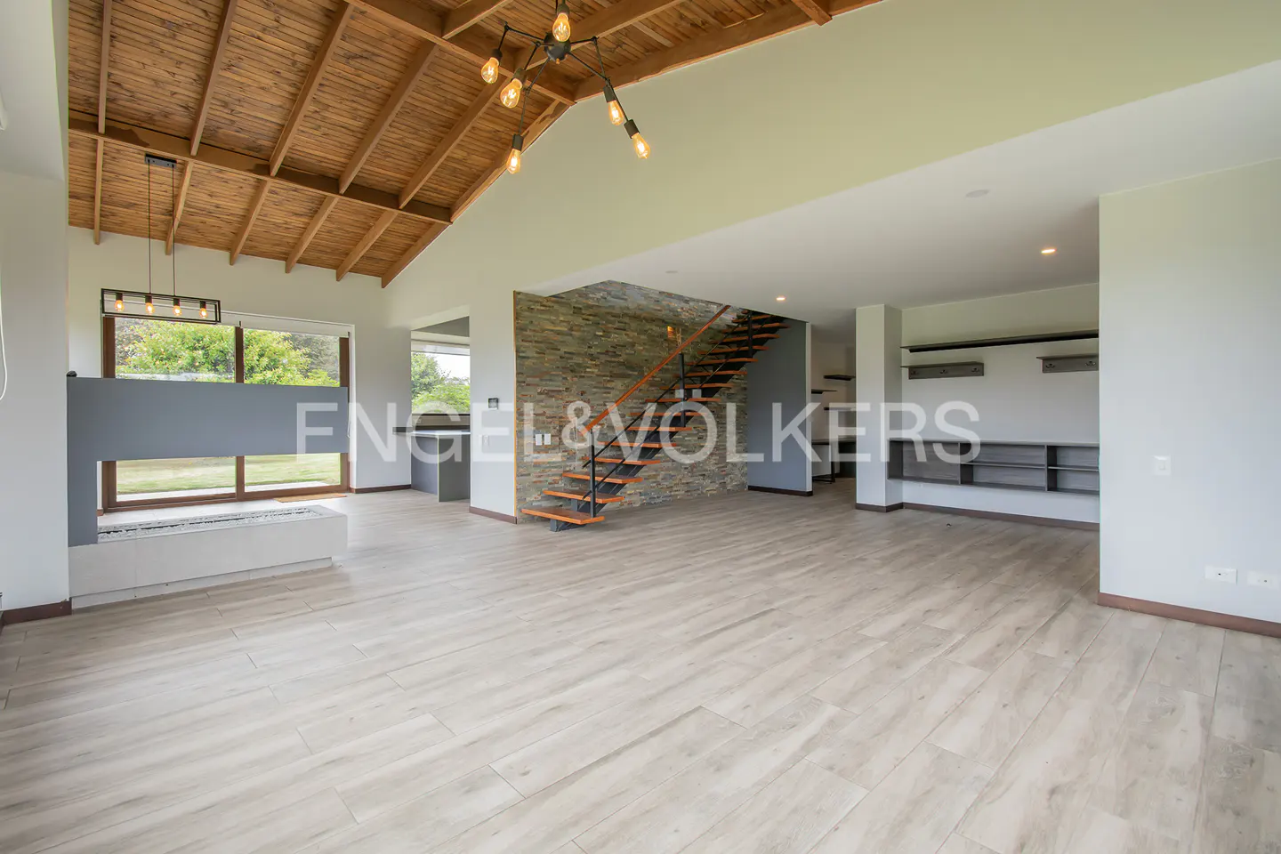 Open-plan living room with light wood floors, stone accent wall, and wooden ceiling. Staircase with wood treads and black metal railing. Modern lighting fixtures.