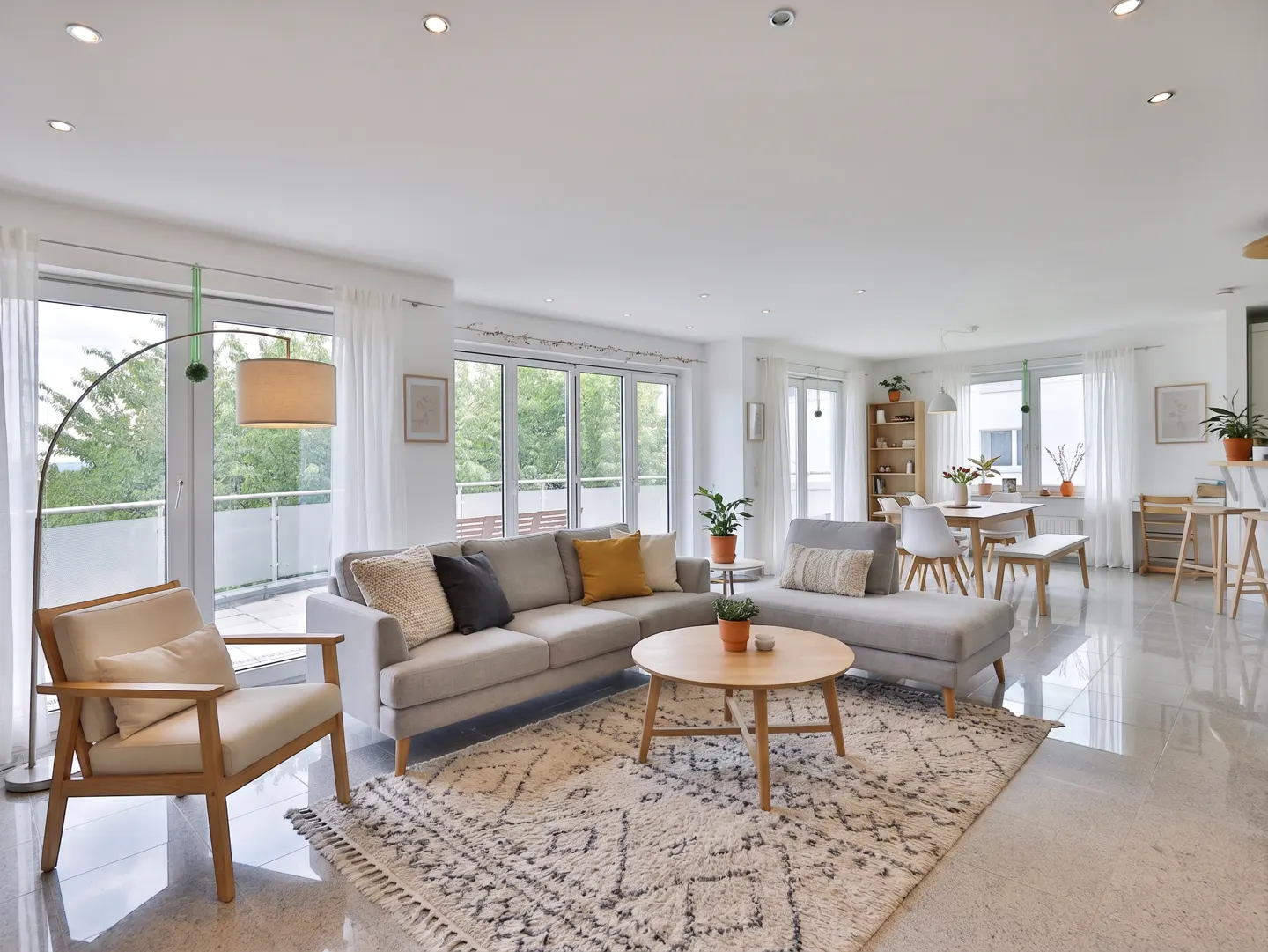 Bright, open-concept living room with a gray sectional sofa, wood chair, and patterned rug. Large windows with white curtains let in natural light.
