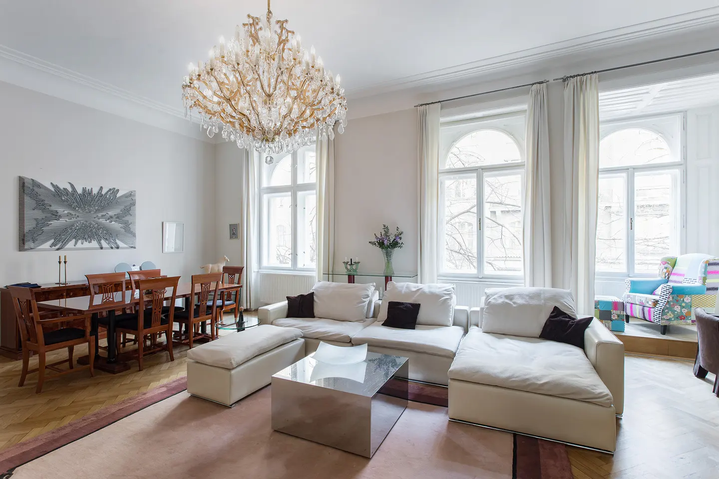Bright living room with a large crystal chandelier, white sofas, a dining table, and tall windows with white curtains.