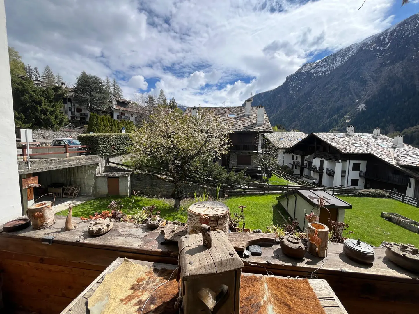 View from a wooden balcony with rustic decor overlooking a green lawn, houses with stone roofs, and a mountain backdrop.